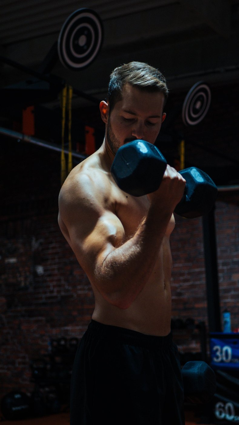 Homme torse nu qui fait un curl avec un haltère dans une salle de sport sombre, devant un mur de briques