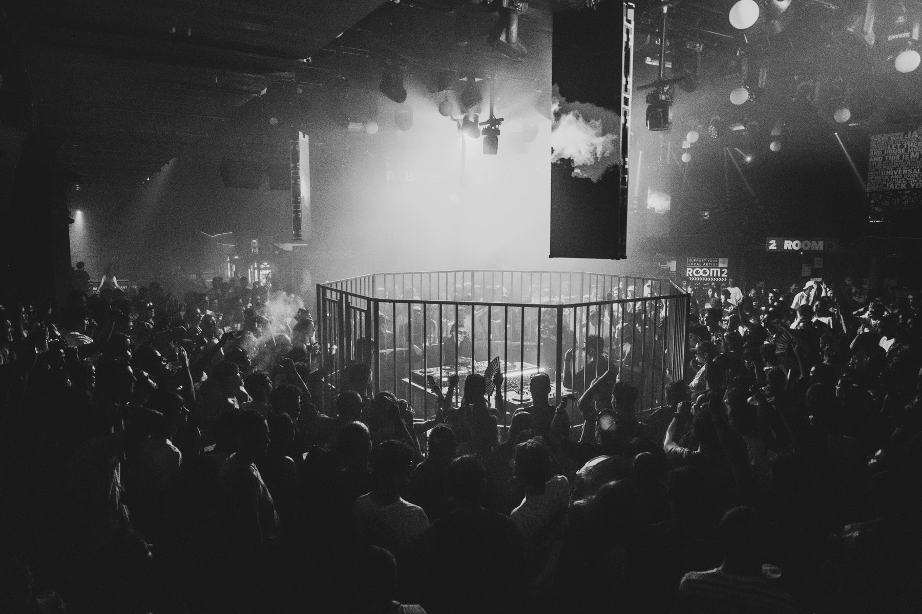 Soirée en club : un DJ dans une cage octogonale au milieu de la foule, dans la fumée, en noir et blanc